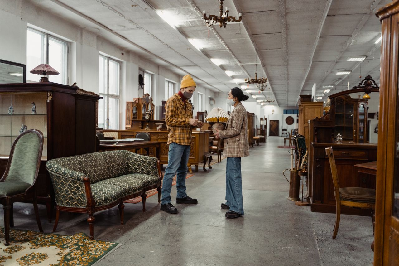 A man and woman wearing masks engage in conversation inside an antique furniture shop.